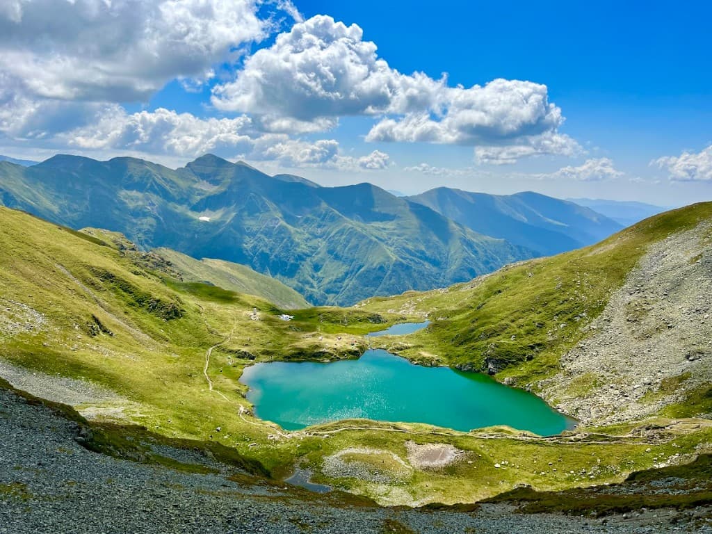 Lac glaciar turcoaz între versanți verzi, poteci de drumeție și creste în Munții Făgăraș, pe cer albastru